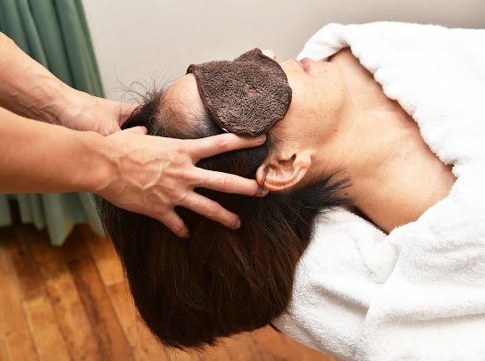 白いタオルをまとい目元にタオルを乗せて横になった女性の頭に、施術者が両手を添えてヘッドマッサージをしている様子
